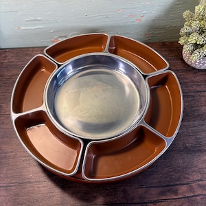 Può includere: Un vassoio da portata vintage con una ciotola centrale in acciaio inossidabile circondata da sei scomparti marroni. Il vassoio ha un design circolare con finiture argentate, perfetto per servire snack o antipasti. Il vassoio è in metallo e plastica.