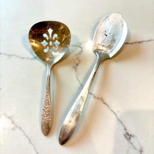 May include: Two antique silver serving spoons. One spoon has a slotted, floral-shaped bowl. The other spoon has a solid, oval-shaped bowl. Both spoons have long, slender handles with floral detailing.
