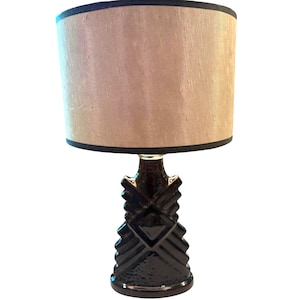 May include: A table lamp featuring a dark brown, glossy, geometric ceramic base with a stepped, angular design. The lamp is topped with a cylindrical lampshade in a light beige, textured fabric with dark brown trim at the top and bottom edges.