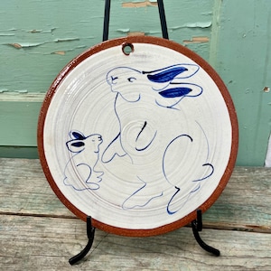 May include: A white ceramic plate with a blue and white illustration of two rabbits. The larger rabbit is standing on its hind legs with its front paws raised. The smaller rabbit is sitting on the ground. The plate has a brown rim and a hole at the top for hanging.
