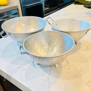 May include: Three vintage aluminium colanders with star-shaped perforations. Each colander has a rounded bowl shape, small feet, and sturdy handles. The colanders are arranged on a white countertop, showcasing their classic design and functionality for kitchen use.