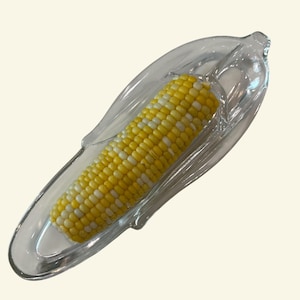 May include: A clear glass corn-on-the-cob dish holds a cooked ear of corn. The corn has yellow and white kernels. The dish is shaped like a corn husk, with a curved, elongated design.