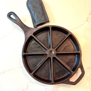 May include: A round black cast iron wedge pan featuring eight triangular compartments and a central circular hole. The pan includes a long handle and a smaller opposite handle, accompanied by a dark grey quilted fabric handle cover.