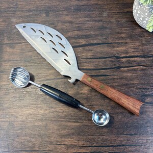 May include: A stainless steel fish-shaped grill knife with a wooden handle and a bottle opener, paired with a melon baller. The knife has a serrated edge and decorative cutouts. The melon baller has a black handle and two stainless steel scoops.