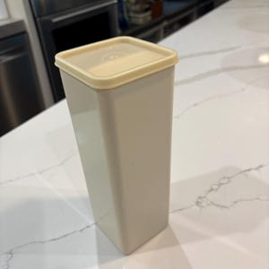 May include: A tall, rectangular, beige container with a square, light yellow lid. The container is made of a smooth, solid material and is designed for storage. The lid fits snugly on top, creating a sealed closure.