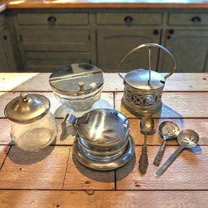 May include: A collection of vintage kitchenware, including glass jars with silver lids, a sugar bowl with a hinged lid, and serving spoons. The items are arranged on a wooden surface, showcasing a classic aesthetic.