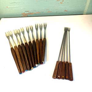 May include: A set of vintage fondue forks with stainless steel tines and dark wooden handles. The handles have colored tips, and a second set of forks is also visible. The forks are arranged on a white surface.