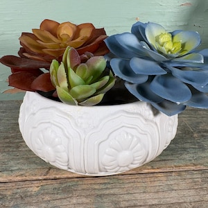 May include: A white ceramic planter with a detailed floral design, filled with a variety of artificial succulents. The succulents are in shades of brown, green, and blue, creating a colorful display. The planter is sitting on a wooden surface.