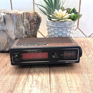 May include: A vintage black and brown Realistic Chronomatic-212 digital alarm clock. The clock displays the time as 12:55 in red digits. The clock has a wood-grain top and is set on a wooden surface.
