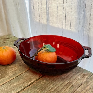 May include: A red glass bowl with two handles and a ribbed design. The bowl contains two oranges.