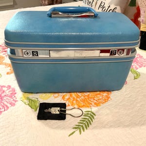 May include: A rectangular light blue hard-shell train case with a matching blue handle and chrome accents. The front features a silver-toned locking mechanism with the letters S and B visible. A single metal key on a ring rests on a small black pouch.