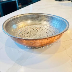 May include: A copper and silver-toned metal colander with a perforated interior. The colander has a wide, shallow bowl shape with a rounded rim. The exterior is copper, and the interior is silver-toned with numerous small holes for draining.