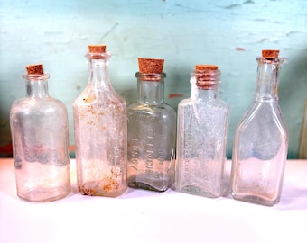 Puede incluir: Cinco botellas de vidrio antiguas con tapones de corcho. Las botellas varían en forma y tamaño, con signos visibles de edad y desgaste. El fondo es una superficie de madera azul claro desgastada.