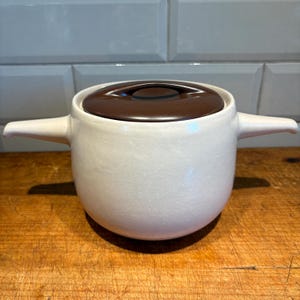 以下が含まれることがあります： 白磁の鍋で、茶色の蓋と2つの取っ手が付いています。鍋は丸みを帯びた形をしていて、滑らかでマットな仕上がりです。