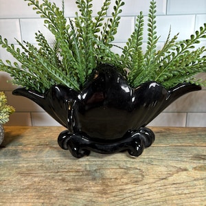 May include: A glossy black ceramic planter with a scalloped rim and a decorative base, filled with vibrant green faux fern stems. The planter sits on a wooden surface, creating a striking contrast.