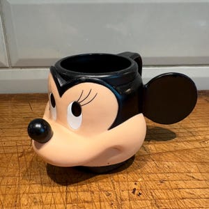May include: A Minnie Mouse-shaped mug with a black handle and ear. The mug features Minnie's face, with a black nose, black hair, and white eyes. The mug is made of ceramic and is a light pink color.
