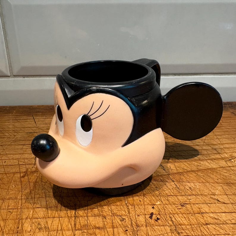 May include: A Minnie Mouse-shaped mug with a black handle and ear. The mug features Minnie's face, with a black nose, black hair, and white eyes. The mug is made of ceramic and is a light pink color.