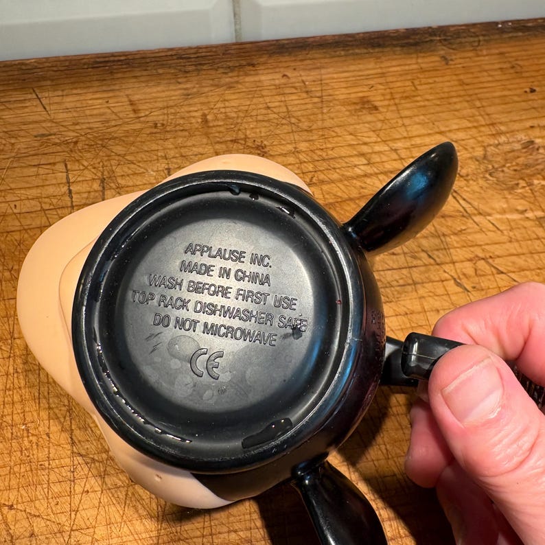 May include: A black and tan character-shaped mug with black handles. The bottom of the mug has printed text that reads "APPLAUSE INC. MADE IN CHINA WASH BEFORE FIRST USE TOP RACK DISHWASHER SAFE DO NOT MICROWAVE CE."
