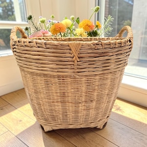 May include: A woven wicker basket with two curved handles, filled with a variety of colorful flowers. The basket is light brown and has a square base. The flowers include orange, yellow, and pink blooms.