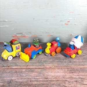 May include: A collection of colorful toy vehicles featuring Sesame Street characters. The set includes a yellow truck, an orange and blue bulldozer, a red train, and a red cement mixer. Each vehicle has rolling wheels.