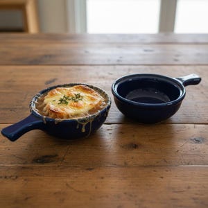 Puede incluir: Dos cuencos de sopa de cerámica azul marino con asas sobre una mesa de madera. Un cuenco está lleno de sopa de cebolla francesa, cubierta con queso derretido y hierbas. El otro cuenco está vacío.