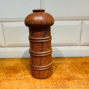 Może przedstawiać: Drewniany solniczka lub pieprzniczka o cylindrycznym korpusie i zaokrąglonym wierzchołku. Solniczka ma ciemnobrązowe usłojenie drewna i jest ozdobiona wieloma drewnianymi paskami.