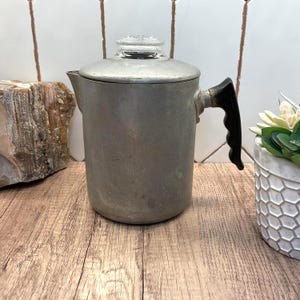 Op de afbeelding: Een vintage aluminium percolator voor op het fornuis met een matte zilveren afwerking, een zwarte hittebestendige handgreep en een doorzichtige glazen knop op het deksel. De cilindrische koffiezetter heeft een eenvoudige tuit.