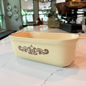 May include: A rectangular ceramic loaf pan in a pale cream color with a subtle crackle glaze finish. The exterior features a centered brown floral scroll pattern. The dish has integrated side handles and a smooth, slightly tapered base design.