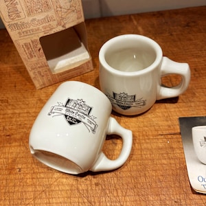 May include: Two white ceramic mugs with the "White Castle" logo in black. The mugs have a rounded shape with a handle. A cardboard box with text is visible in the background. The mugs are on a wooden surface.