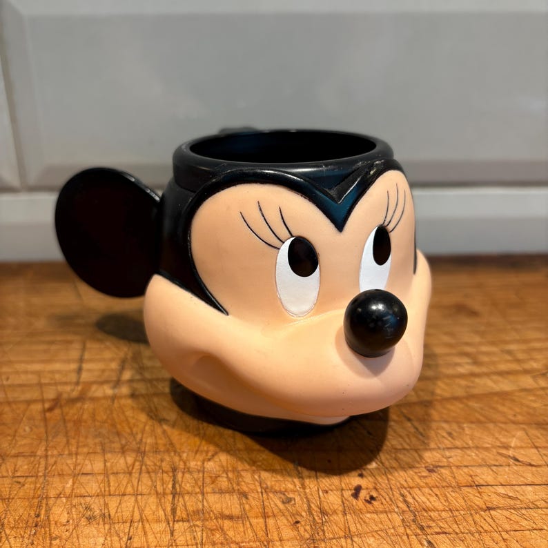 May include: A Minnie Mouse-shaped mug with a black handle and black interior. The mug features Minnie's face, with a black nose, eyelashes, and ears. The mug is made of a light pink material.