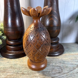 May include: A carved wooden pineapple-shaped ornament with a flower-like top. The brown ornament has a diamond pattern and small holes. Two other dark brown wooden objects are in the background.