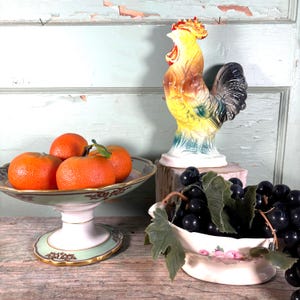 Puede incluir: Una colorida estatua de gallo de cerámica se encuentra sobre un bloque de madera junto a un frutero decorativo lleno de naranjas. También hay un plato pequeño con uvas y un cuenco decorativo con naranjas. El gallo es amarillo, naranja y negro.