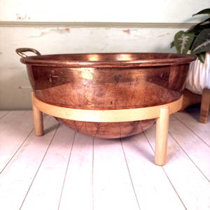 Puede incluir: Un cuenco de cobre pulido, apoyado sobre un soporte de madera clara. El cuenco tiene forma redondeada con un pequeño asa en un lado. El soporte tiene tres patas de madera y un soporte curvo.