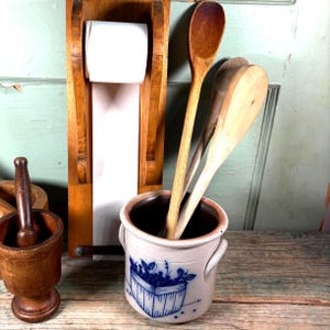 May include: A collection of kitchen items, including wooden spoons, a ceramic utensil holder with a blue berry basket design, a wooden paper towel holder, and a mortar and pestle.
