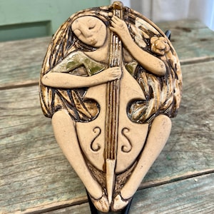 May include: A ceramic wall sculpture of a person playing a cello. The sculpture is painted in shades of brown, green, and white.