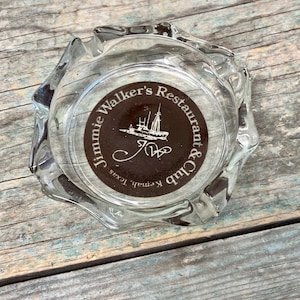 May include: A clear glass ashtray with a brown circular sticker that reads "Jimmie Walker's Restaurant & Club, Kennith Texas" with a white ship and the initials "JW" in a cursive font.