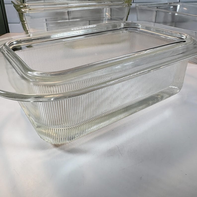 May include: Clear glass rectangular baking dishes with lids. The dishes have a ribbed design on the sides and a smooth, clear lid. The image shows several dishes stacked together, suggesting a set of bakeware.