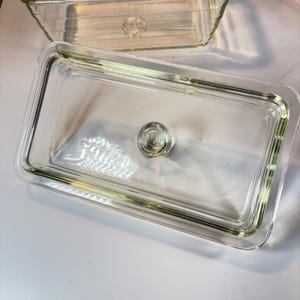 May include: Clear rectangular glass dish with a matching lid. The lid has a small, round handle in the center. The dish is designed for food storage and is made of transparent glass.