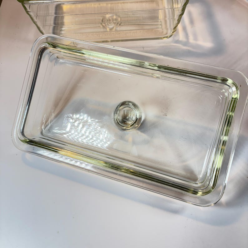 May include: Clear rectangular glass dish with a matching lid. The lid has a small, round handle in the center. The dish is designed for food storage and is made of transparent glass.