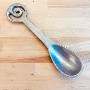 Puede incluir: Una cuchara de metal plateado con un mango largo y recto que termina en un diseño circular de espiral calado. El cuenco de la cuchara es de forma ovalada y lisa, con un acabado metálico uniforme en todo el objeto.
