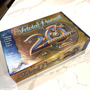 May include: A gold and blue Trivial Pursuit 20th Anniversary Edition board game box. The box features the game's logo and the text "20th Anniversary Edition". The box is on a white marble surface.