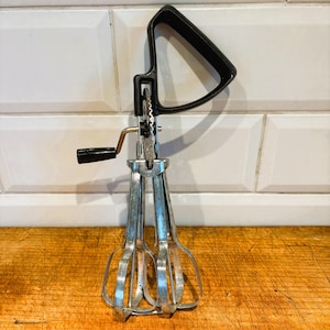 Könnte beinhalten: Ein vintage Handrührgerät mit schwarzem Griff und drei silbernen Rührbesen. Das Rührgerät befindet sich auf einer Holzoberfläche.