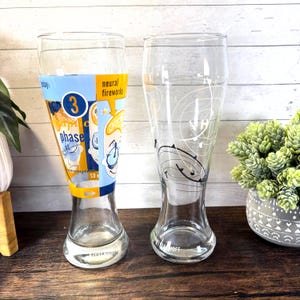 May include: Two clear glass beer glasses. One glass has a blue, yellow, and white graphic with the number 3 and the words "phase" and "neural fireworks". The other glass has a white graphic design.
