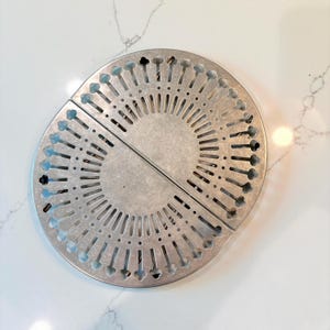 Peut inclure: Un dessous de plat circulaire en métal argenté composé de deux moitiés semi-circulaires identiques. La surface présente un motif radial de fentes allongées et de petites découpes en forme de trèfle sur le bord extérieur, créant un design perforé.