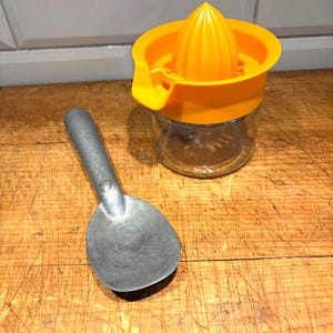 May include: A vintage kitchen set featuring a metal ice cream scoop and a yellow citrus juicer with a clear glass container. The juicer has a ribbed cone and a pouring spout. The items are on a wooden surface.