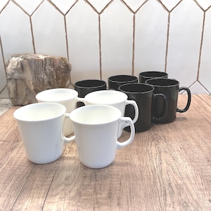 May include: A selection of white and black ceramic mugs with curved handles. The mugs are displayed on a wooden surface, with a white tile backsplash. These mugs are suitable for hot drinks such as tea or coffee.
