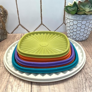 May include: A stack of five square plastic lids in descending sizes, featuring a sunburst pattern. The colors from bottom to top are off-white, teal, purple, orange, and lime green. Each lid has a raised, ridged edge and a central circular detail.