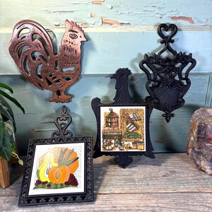 May include: A collection of vintage kitchen decor items. Includes a copper rooster wall hanging, two black trivets with decorative tile inserts, and a black cast iron trivet with a heart-shaped handle. The tiles feature fruit and a vintage scene.
