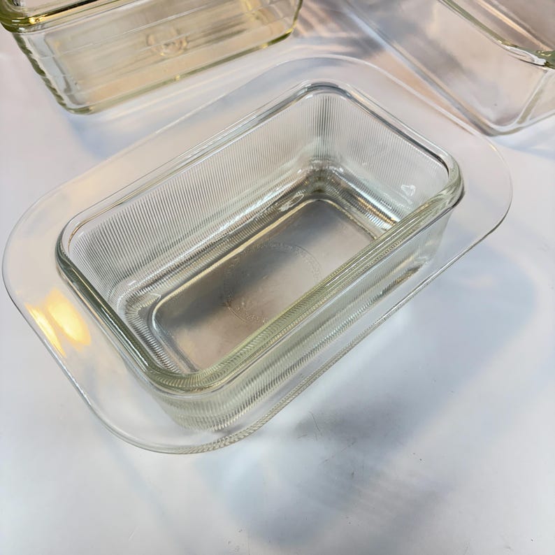 May include: Clear glass loaf pan with a ribbed texture, resting on a clear glass tray. The pan is rectangular with rounded corners. Additional clear glass loaf pans are visible in the background.