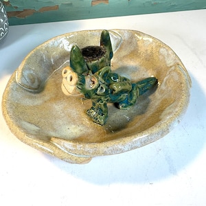 Pode incluir: Um cinzeiro de cerâmica feito à mão com um design caprichoso. O cinzeiro apresenta um prato oval castanho claro com uma borda elevada. Uma figura de dragão verde e bege repousa no interior, com um tronco de árvore castanho escuro. O cinzeiro é uma peça decorativa única.
